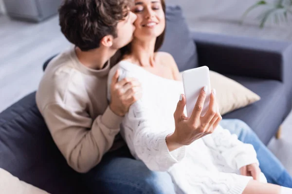 Smartphone in hand of smiling woman taking selfie with boyfriend on blurred background — Stock Photo