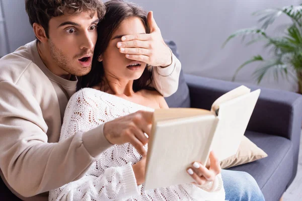 Geschockter Mann verdeckt Freundin Augen beim Lesen von Buch auf verschwommenem Vordergrund — Stockfoto