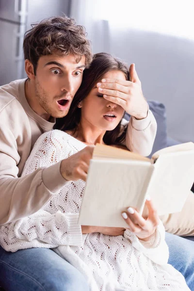 Excited man covering girlfriend eyes while reading book on blurred foreground at home — Stock Photo