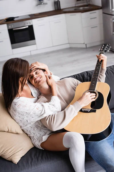 Mulher abraçando namorado sorridente com guitarra acústica no sofá — Fotografia de Stock