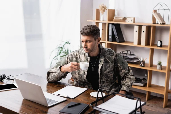 Militar en uniforme sosteniendo taza mientras mira el portátil - foto de stock