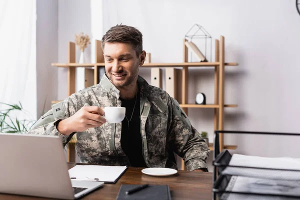 Happy military man in uniform holding cup while looking at laptop near document tray on blurred foreground — Stock Photo