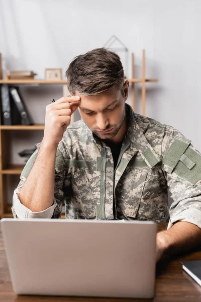 Soldado pensativo en uniforme sentado en el escritorio y usando el ordenador portátil - foto de stock