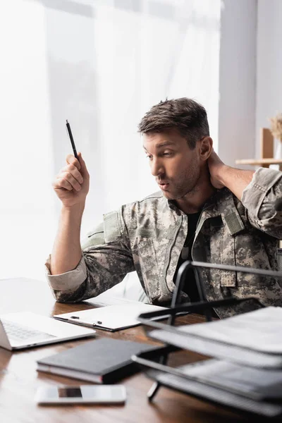 Soldato sorpreso in uniforme che tiene penna vicino al computer portatile con smartphone in primo piano sfocato — Foto stock