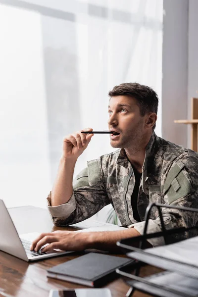 Soldato pensieroso in penna tenuta uniforme vicino al computer portatile con smartphone in primo piano sfocato — Foto stock