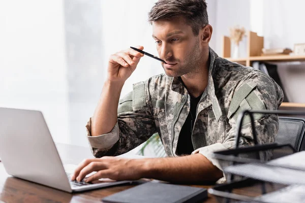 Pensive soldier in uniform holding pen and using laptop near document tray on blurred foreground — Stock Photo