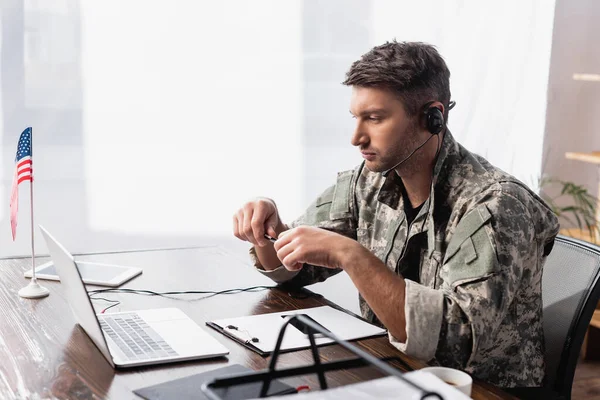 Homem militar patriótico em fone de ouvido segurando caneta e olhando para laptop perto da bandeira americana — Fotografia de Stock