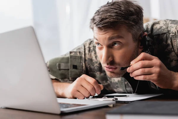 Homem militar em uniforme sussurrando em microfone no fone de ouvido perto do laptop — Fotografia de Stock