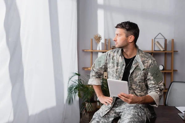 Soldado en uniforme militar sosteniendo tableta digital y mirando hacia otro lado en la oficina - foto de stock