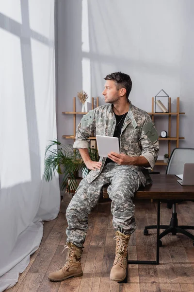 Солдат у військовій формі тримає цифровий планшет біля ноутбука на столі — стокове фото