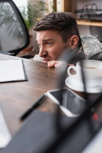 Homem militar descontente escondido debaixo da mesa com xícara de café e smartphone — Fotografia de Stock