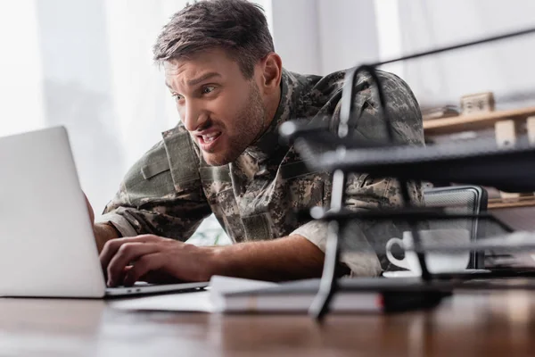 Irritated military man typing on laptop keyboard near document tray on blurred foreground — Stock Photo