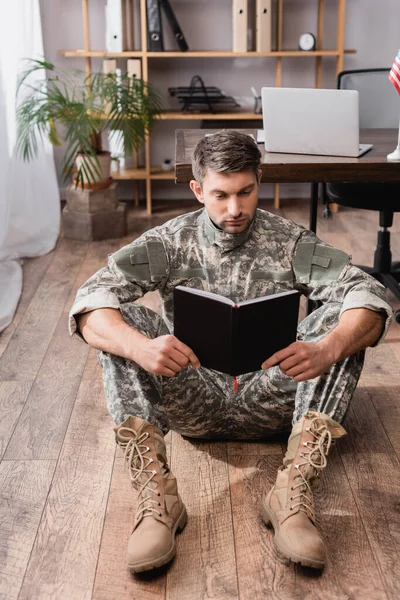 Militar enfocado sosteniendo libro de copias mientras está sentado en el piso cerca del escritorio - foto de stock