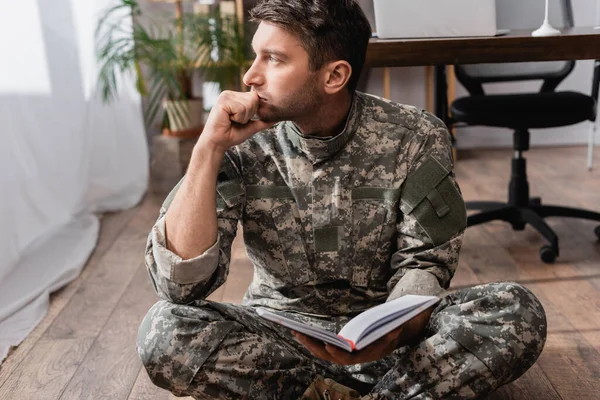 Militar pensativo sosteniendo libro de copia mientras está sentado en el piso cerca del escritorio - foto de stock
