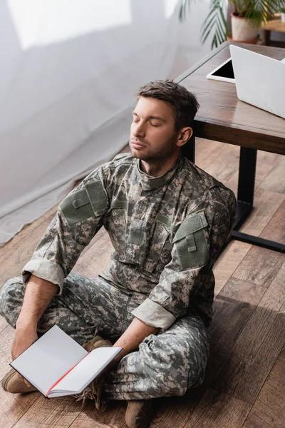 Militar cansado sosteniendo libro de copias en blanco mientras está sentado en el piso cerca del escritorio - foto de stock