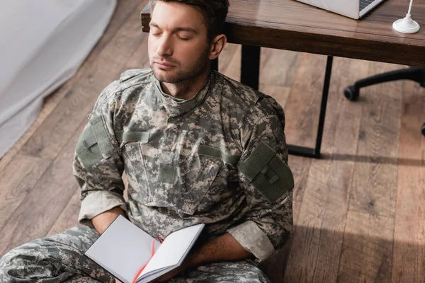 Militar sosteniendo libro de copias en blanco mientras está sentado en el piso cerca del escritorio - foto de stock