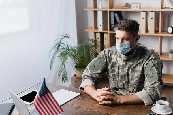 Militaire en uniforme et masque médical assis les mains serrées au bureau avec drapeau américain — Photo de stock
