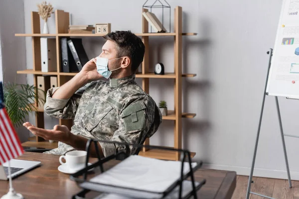 Militaire en uniforme et masque médical parlant sur smartphone près du drapeau américain sur le bureau — Photo de stock