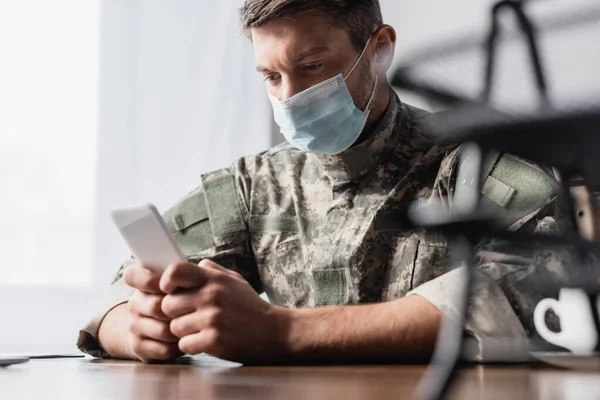 Teléfono inteligente en las manos en el militar en uniforme y máscara médica cerca de bandeja documental en primer plano borrosa - foto de stock