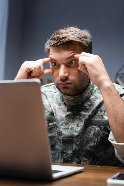 Измученный военный в форме смотрит на ноутбук на размытом переднем плане — стоковое фото