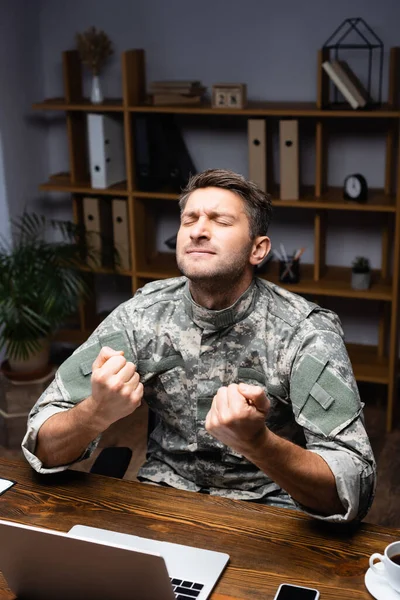 Homme militaire excité en uniforme assis avec les poings serrés tout en se réjouissant près de l'ordinateur portable et smartphone sur le bureau — Photo de stock