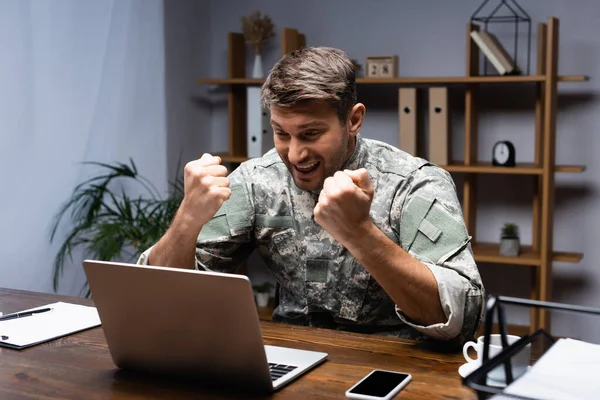 Excited military man sitting with clenched fists while rejoicing near laptop and smartphone on desk — Stock Photo