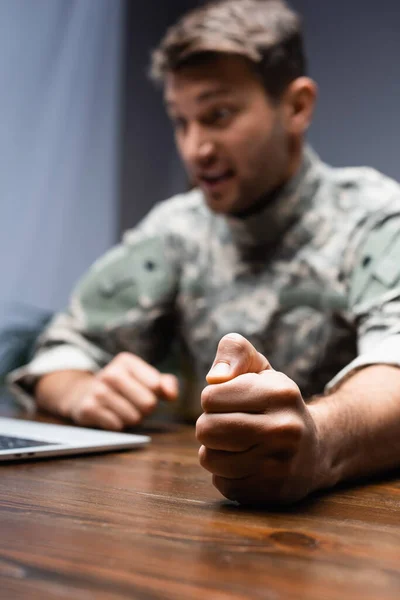 Militar en uniforme sentado con el puño cerrado cerca de la computadora portátil sobre fondo borroso - foto de stock