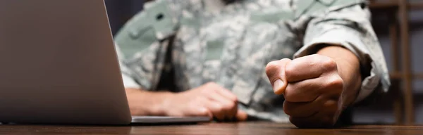 Vista recortada del militar en uniforme sentado con el puño cerrado cerca de la computadora portátil, pancarta - foto de stock