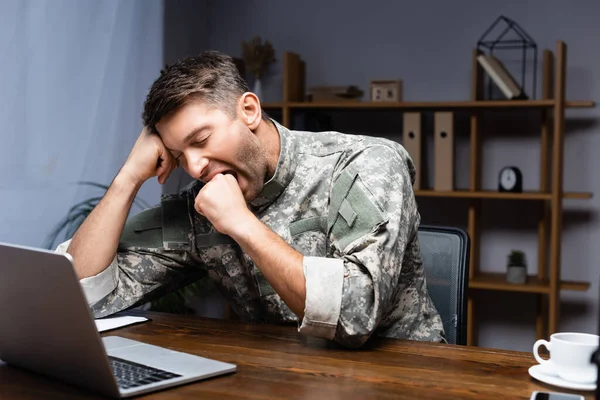 Hombre militar cansado en uniforme sentado con el puño cerrado mientras bosteza cerca de la computadora portátil - foto de stock