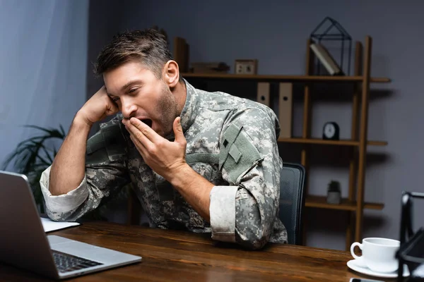 Tired military man in uniform covering mouth while yawning near laptop — Stock Photo