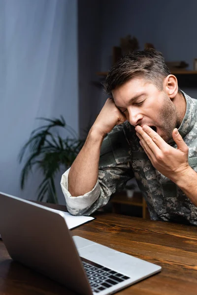 Erschöpfter Soldat in Uniform verdeckt Mund, während er neben Laptop gähnt — Stockfoto