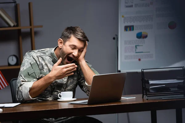 Сонный военный в форме, прикрывающий рот, зевая возле ноутбука. — стоковое фото