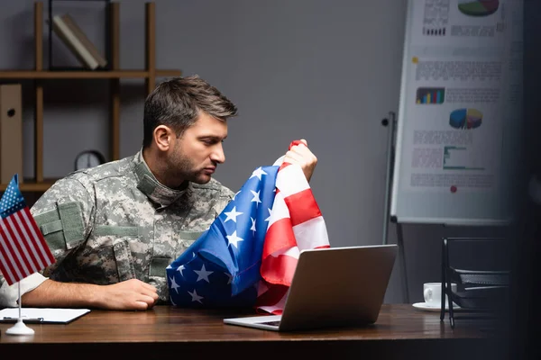 Сумний військовий чоловік у формі тримає прапор Америки біля ноутбука — стокове фото