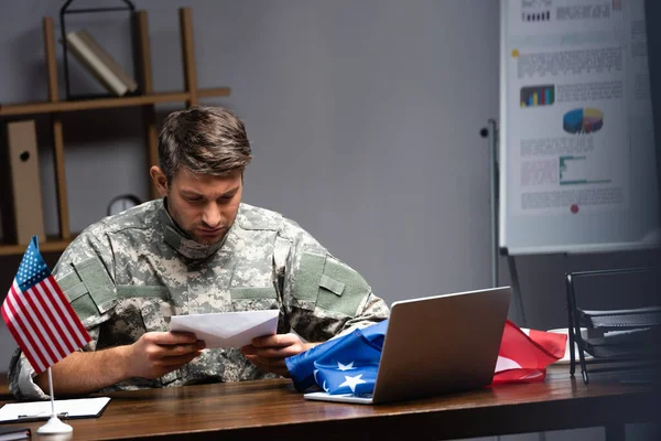Сумний військовий чоловік у формі, що тримає конверт біля ноутбука та американських прапорів — стокове фото