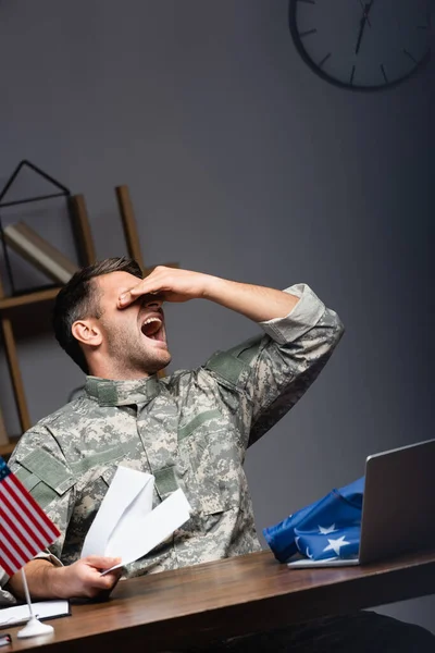 Homem militar zangado em uniforme cobrindo olhos e segurando carta enquanto gritando perto de laptop e bandeiras americanas — Fotografia de Stock