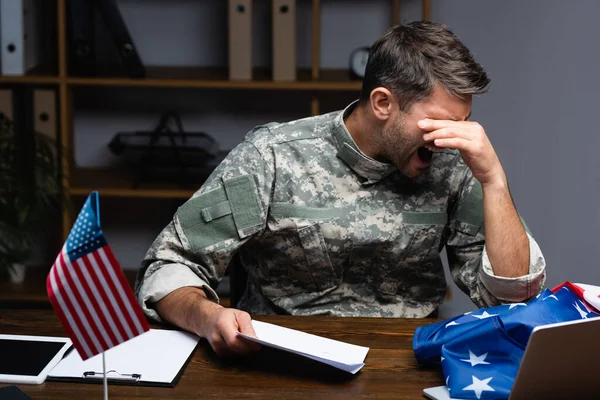Molesto militar en uniforme cubriendo los ojos y sosteniendo la carta mientras grita cerca de gadgets y bandera americana - foto de stock