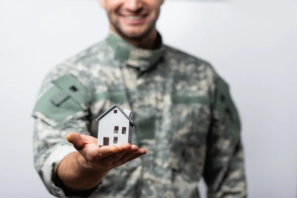 Modelo de casa en la mano del hombre militar feliz en uniforme sobre fondo borroso aislado en blanco - foto de stock