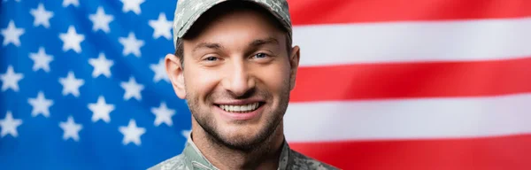 Feliz militar en uniforme y gorra sonriendo cerca de bandera americana sobre fondo borroso, bandera - foto de stock
