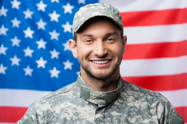 Feliz militar en uniforme y gorra sonriendo cerca de bandera americana sobre fondo borroso - foto de stock