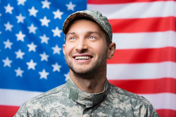 Fröhlicher Soldat in Uniform und Mütze, der neben der amerikanischen Flagge auf verschwommenem Hintergrund lächelt — Stockfoto