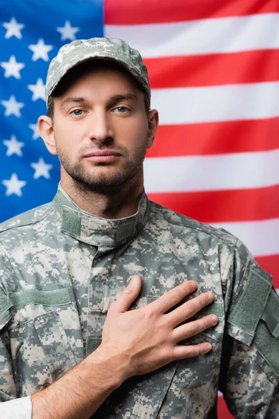 Patriottico militare in uniforme giuramento fedeltà vicino bandiera americana su sfondo sfocato — Foto stock