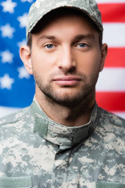 Military man in uniform and cap looking at camera near american flag on blurred background — Stock Photo