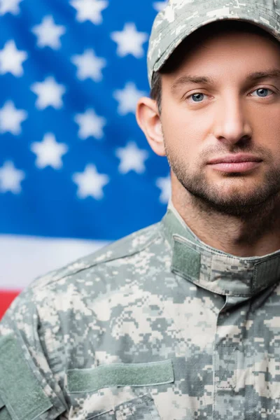 Homem militar de uniforme olhando para a câmera perto da bandeira americana em fundo embaçado — Fotografia de Stock