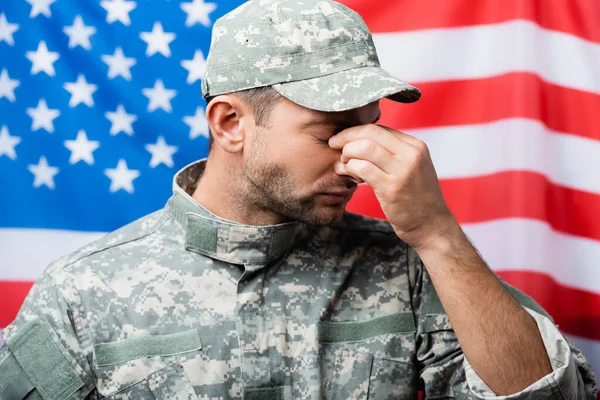 Triste uomo militare in uniforme e tappo asciugare le lacrime mentre piange vicino bandiera americana su sfondo sfocato — Foto stock