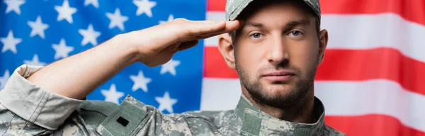 Patriotic military man in uniform and cap giving salute near american flag on blurred background, banner — Stock Photo