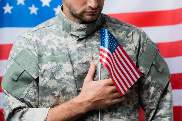 Обрізаний вид патріотичного військового в уніформі, що тримає маленький американський прапор на розмитому тлі — стокове фото