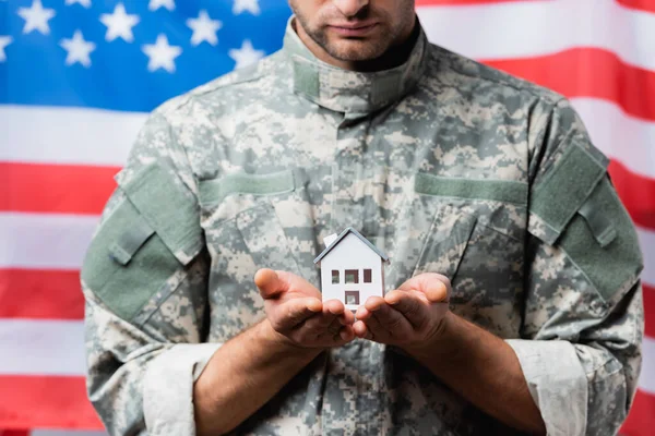 Патриотический военный в форме держит модель дома рядом с американским флагом на размытом фоне — стоковое фото