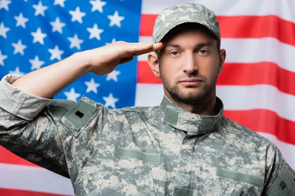 Militar patriótico en uniforme y gorra dando saludo cerca de bandera americana sobre fondo borroso - foto de stock