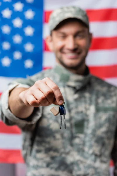 Keys in hand of patriotic military man in uniform near american flag on blurred background — Stock Photo