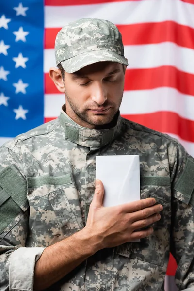 Homme militaire patriotique en uniforme et casquette tenant enveloppe près du drapeau américain sur fond flou — Photo de stock